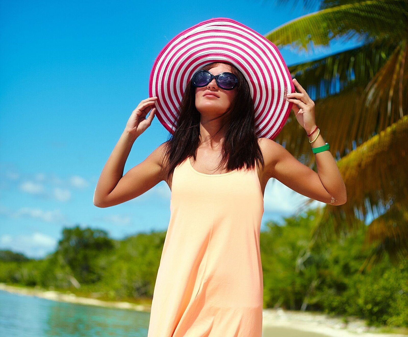 Hot beautiful woman in colorful sunhat and dress walking near beach ocean on hot summer day near palm